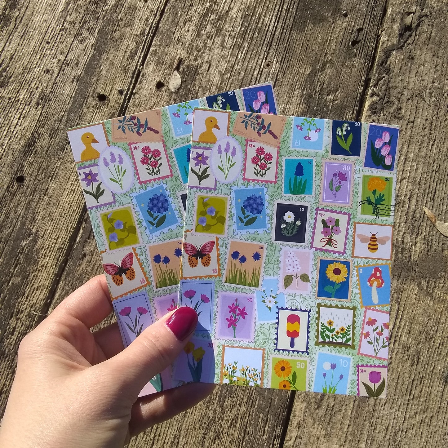 Hand holding a small stack of colorful postcards with nature-themed designs on a wooden surface.