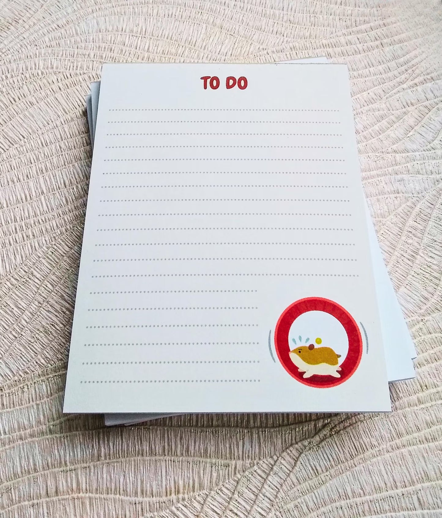 Picture of a white notepad on a textured surface. It says 'To Do' at the top in red. It is lined with dotted lines and has an illustration of a hamster running in a wheel in the bottom right corner.