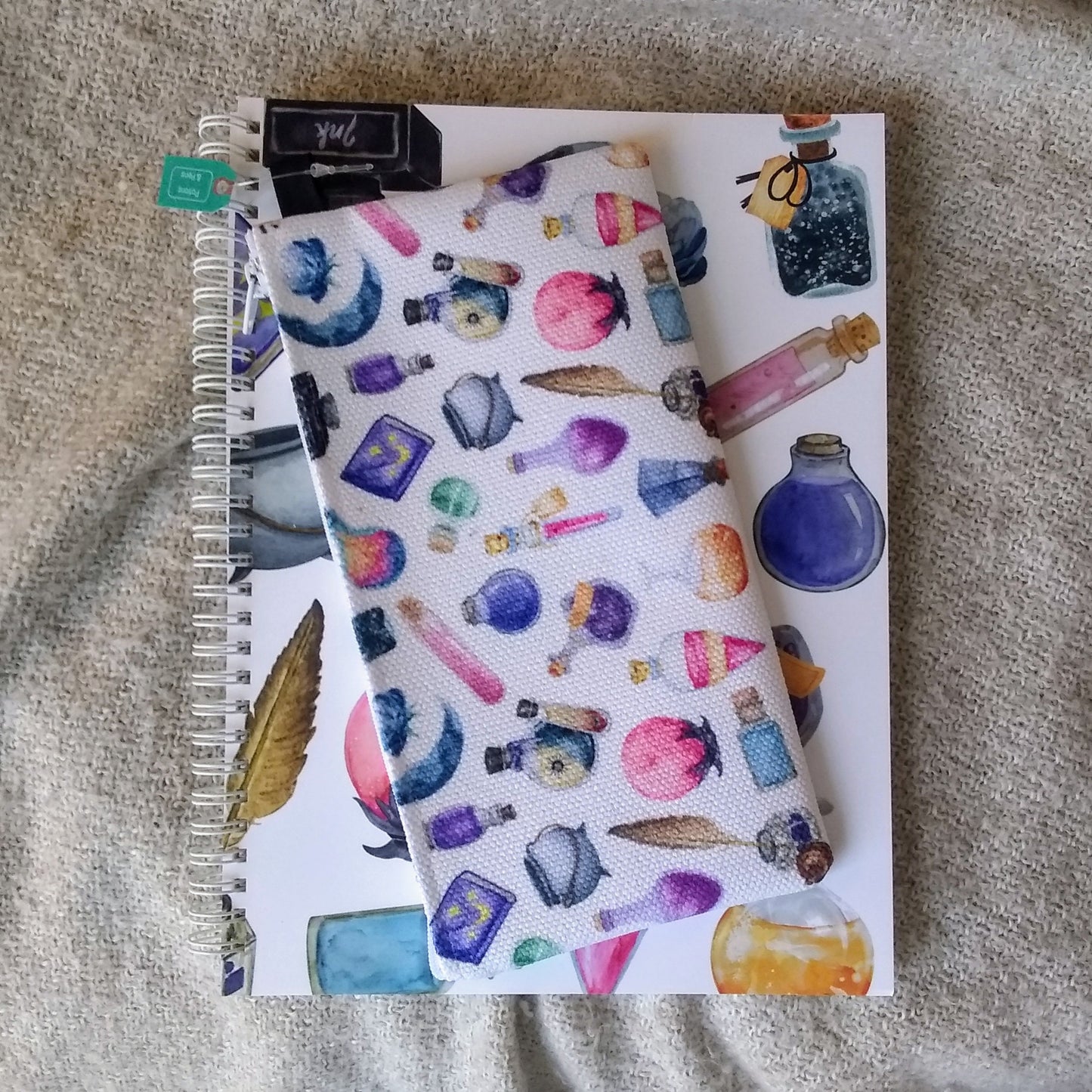 Notebook with colorful potion bottle pattern on a textured surface. On top is a rectangular cloth pencil case with the same design.
