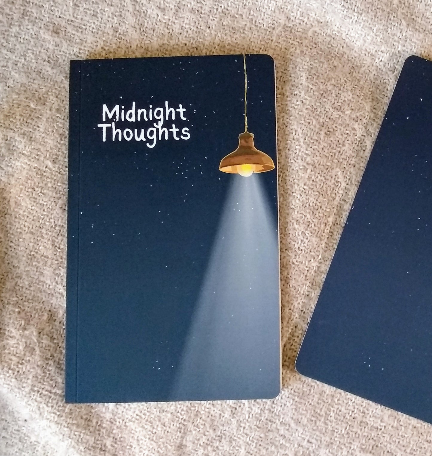 Notebook with 'Midnight Thoughts' on the cover, illustration of a starry night sky illuminated by a hanging lamp, sitting on a textured surface