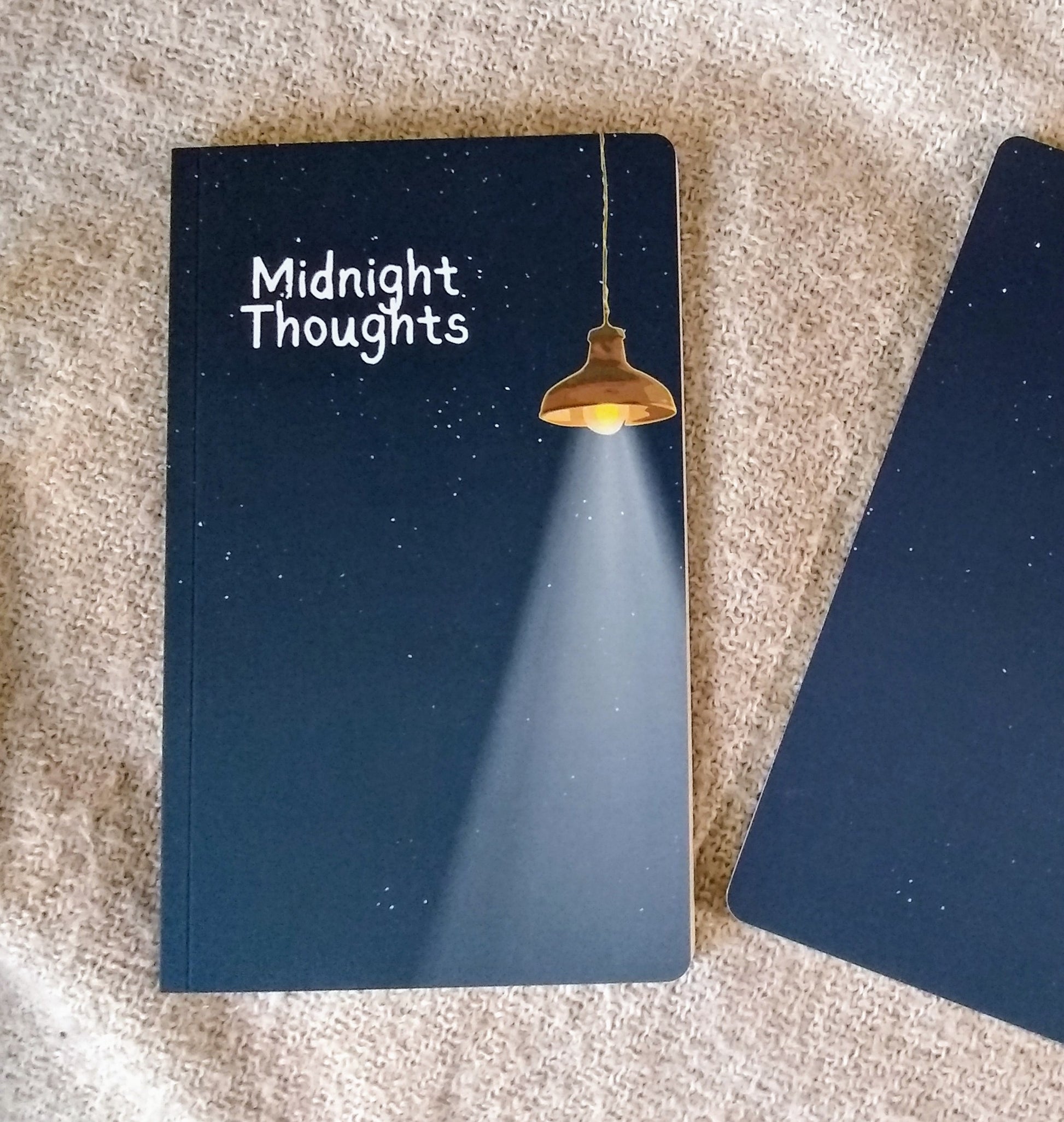 Notebook with 'Midnight Thoughts' on the cover, illustration of a starry night sky illuminated by a hanging lamp, sitting on a textured surface