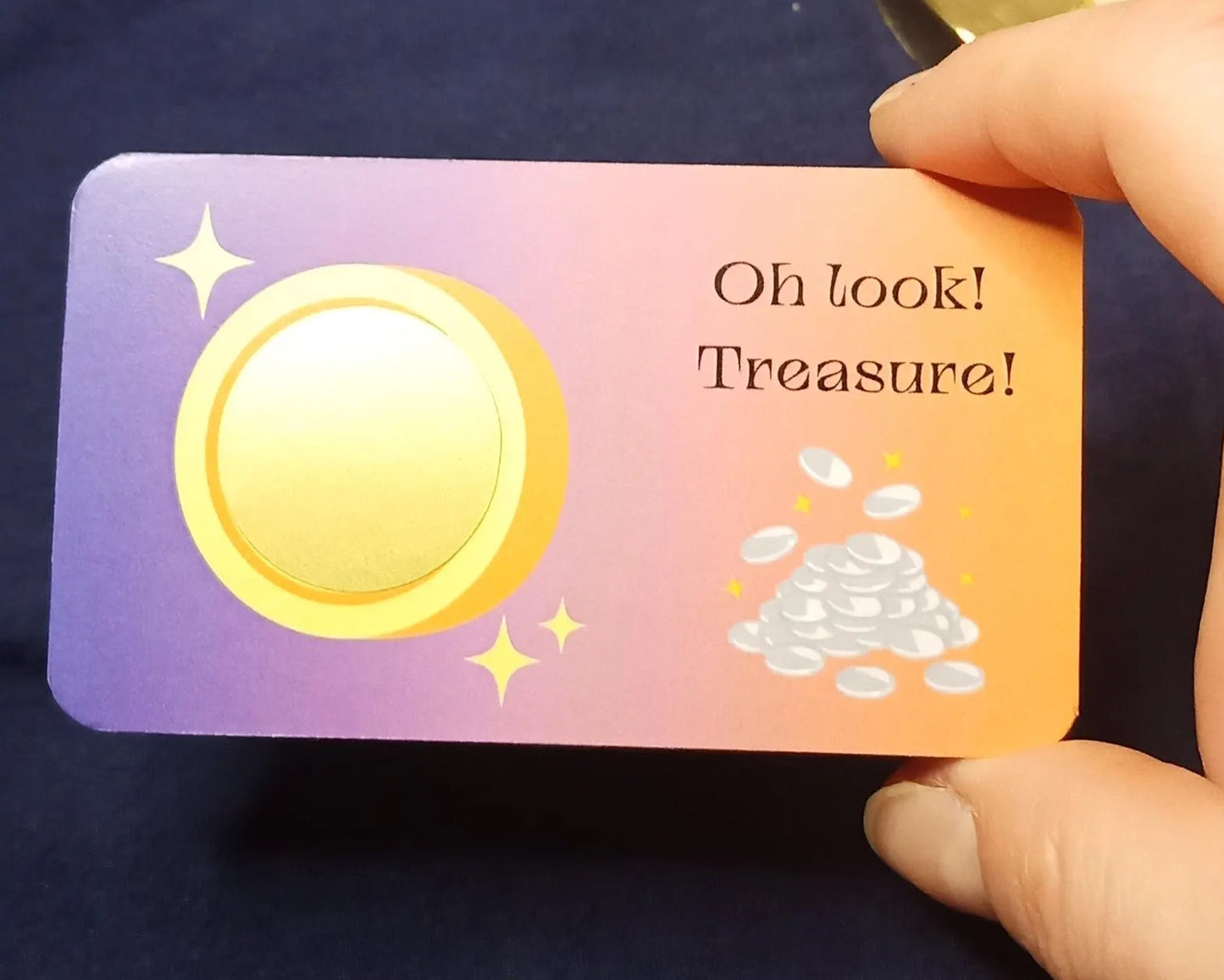 Card with a gold coin and text 'Oh look! Treasure!' held by a hand against a dark background.