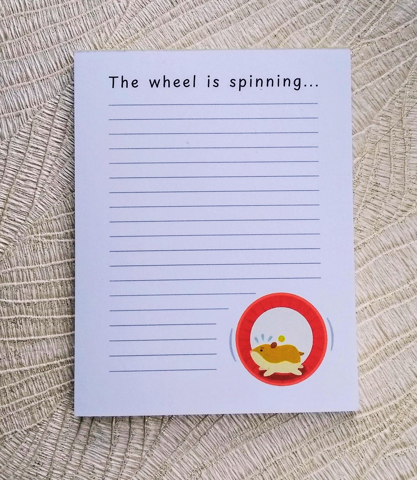White notepad with lines down the front, and an illustration of a hamster running in a wheel in the bottom right corner. At the top it says "The wheel is spinning..." in black font.