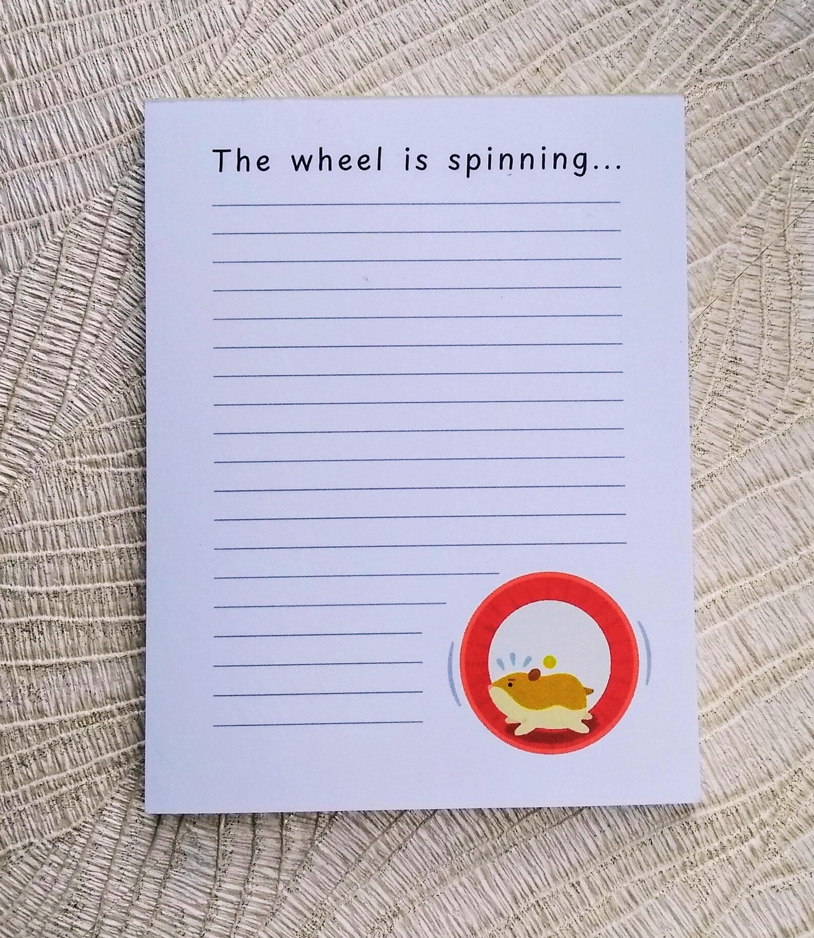 White notepad with lines down the front, and an illustration of a hamster running in a wheel in the bottom right corner. At the top it says "The wheel is spinning..." in black font.