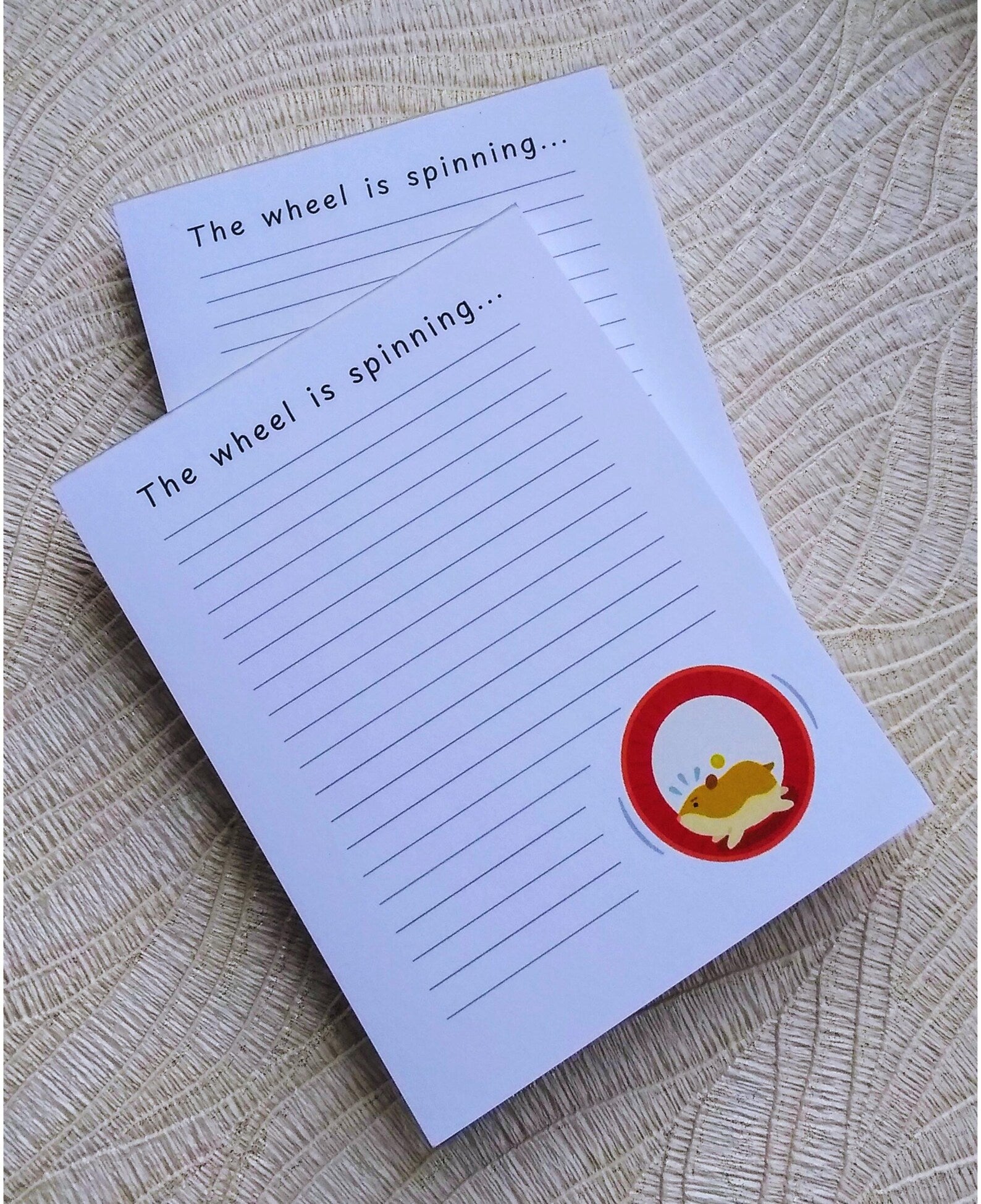 Picture of two notepads with one on top of another. The notepads have lines down the front and an illustration of a hamster running in a wheel in the bottom right corner. At the top it says "The wheel is spinning..." in black font.
