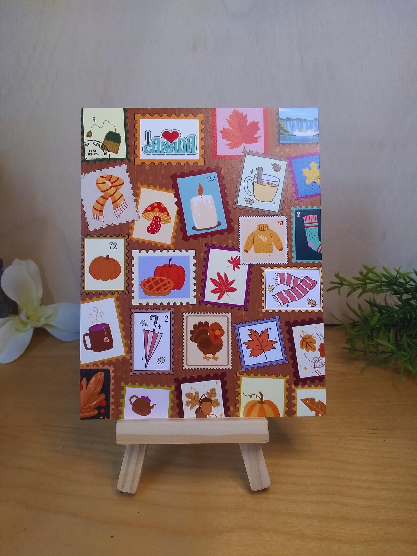 Picture of a postcard standing vertically. It has a background of wood with a collage of illustrated cartoon stamps in an autumn or fall theme. They show seasonal items like a scarves, leaves, a turkey, a sweater, pumpkins, a candle, and one stamp at the top that says 'I heart Canada".