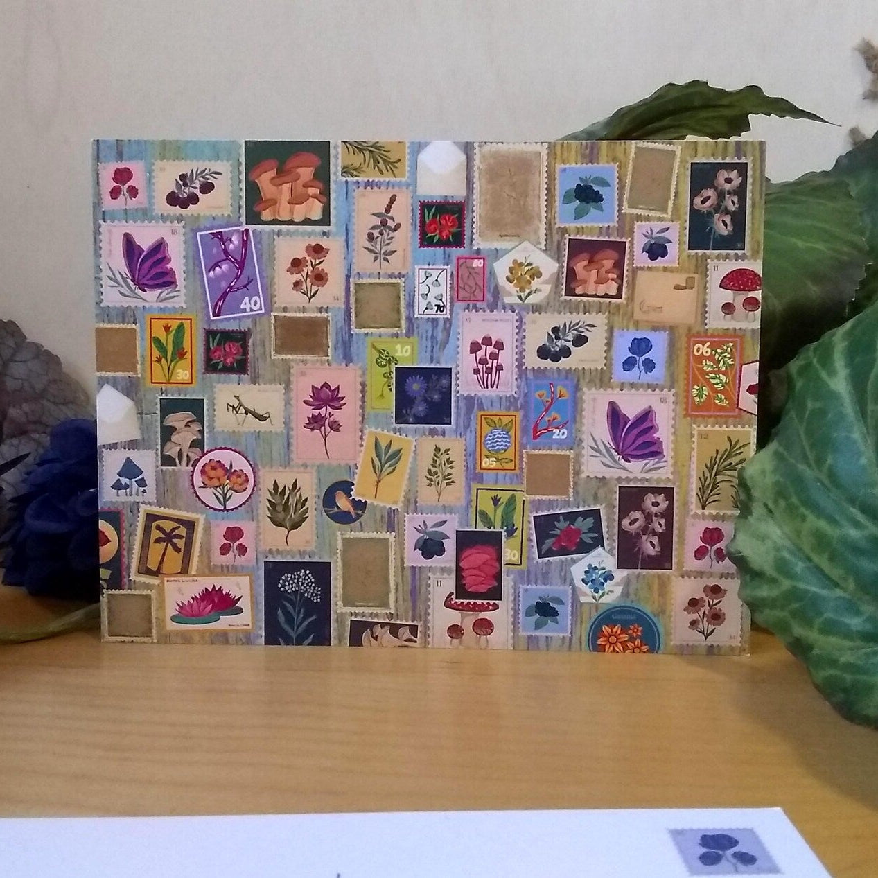 Postcard with colorful stamp-like design on a wooden surface with plants in the background