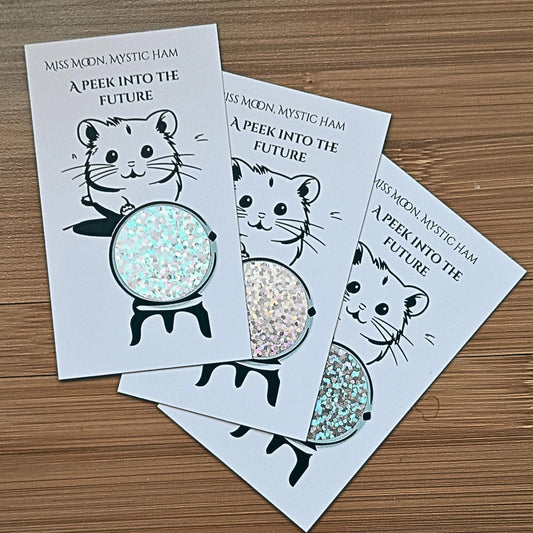 Picture of 3 white rectangular cards fanned out atop a brown surface. The cards are the same and show an illustration of a hamster holding out one paw to a crystal ball. The crystal ball is shiny with a glittery look to it. The top of the card says 'Miss Moon, Mystic Ham. A peek into the future'